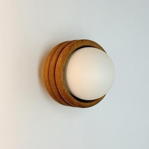 Puede incluir: Aplique de pared moderno con una pantalla esférica blanca rodeada por tres anillos concéntricos de madera. La madera tiene un tono cálido y natural. Montado en una pared blanca.