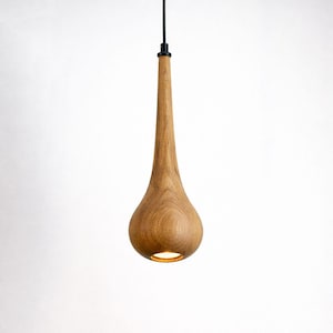 May include: A wooden pendant light fixture with a teardrop-shaped shade. The light fixture is made of light-colored wood and hangs from a black cord. The light is on, illuminating the bottom of the shade.