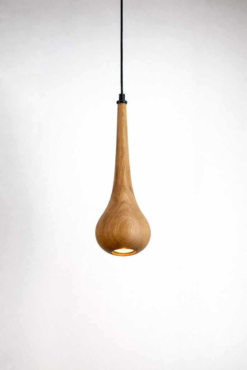 May include: A wooden pendant light fixture with a teardrop-shaped shade. The light fixture is made of light-colored wood and hangs from a black cord. The light is on, illuminating the bottom of the shade.