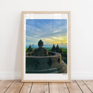 Könnte beinhalten: Ein gerahmter vertikaler Druck mit einer steinernen Buddha-Statue, die in einer Stupa in Borobudur sitzt und eine Landschaft bei Sonnenaufgang mit blauem und gelbem Himmel überblickt. Der Text "Lakhna Raja" steht unten rechts im Bild.