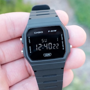 Puede incluir: Un reloj de pulsera digital negro con esfera rectangular y correa de resina negra a juego. La pantalla muestra el texto CASIO F-91W, el día SU, la fecha 1, la hora 12:40:22 y las letras WR. Tiene botones laterales de color plateado.