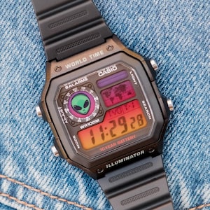 May include: Black digital watch with a rectangular face, featuring a green alien graphic and the text "CASIO". The watch displays the time 11:29:28 on a digital screen. The watchband is black and the watch is on a denim background.