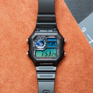 Puede incluir: Reloj digital Casio negro con esfera rectangular y correa negra. El reloj muestra la hora, la fecha y las funciones de hora mundial. El reloj tiene el texto "WORLD TIME", "CASIO" e "ILLUMINATOR".