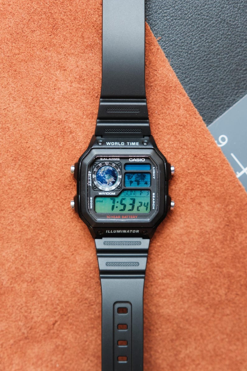 Puede incluir: Reloj digital Casio negro con esfera rectangular y correa negra. El reloj muestra la hora, la fecha y las funciones de hora mundial. El reloj tiene el texto "WORLD TIME", "CASIO" e "ILLUMINATOR".