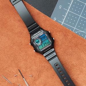 Peut inclure: Montre num&eacute;rique Casio noire avec un cadran rectangulaire affichant l'heure et la date. Le bracelet noir est pos&eacute; sur une surface en cuir marron. Un petit tournevis et des outils de r&eacute;paration de montre sont &agrave; proximit&eacute;.
