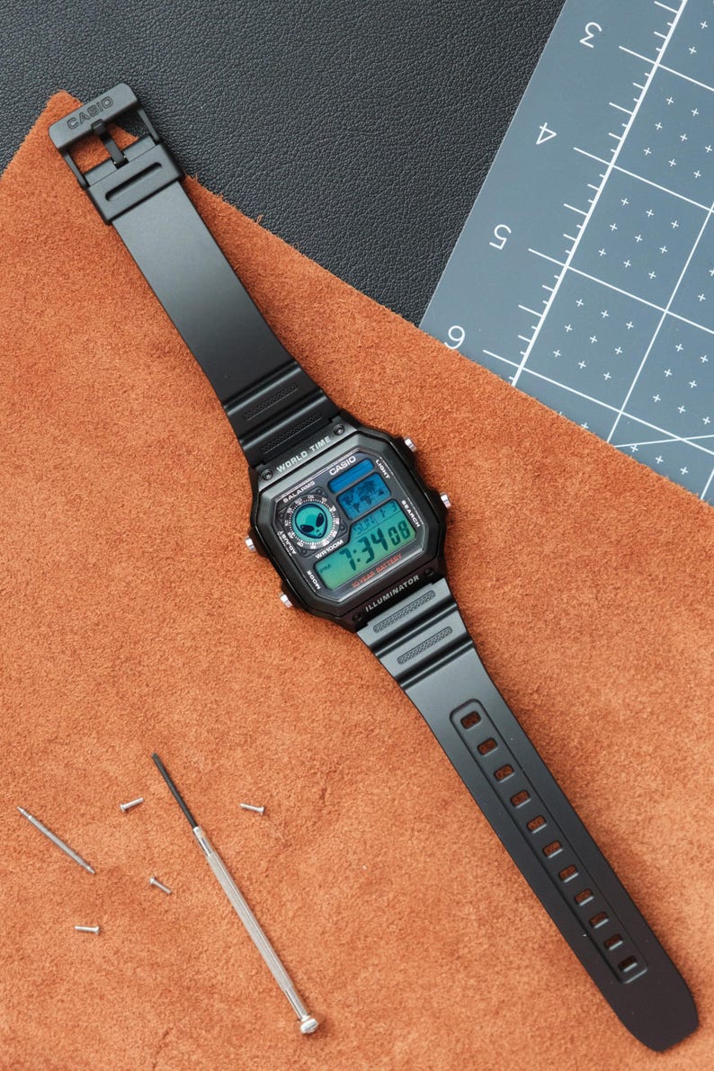 Peut inclure: Montre num&eacute;rique Casio noire avec un cadran rectangulaire affichant l'heure et la date. Le bracelet noir est pos&eacute; sur une surface en cuir marron. Un petit tournevis et des outils de r&eacute;paration de montre sont &agrave; proximit&eacute;.