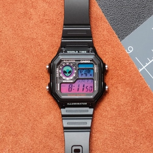 Peut inclure: Montre num&eacute;rique noire avec un cadran carr&eacute; et un bracelet noir. L'&eacute;cran affiche l'heure, la date et d'autres fonctions. La montre porte l'inscription "WORLD TIME" et "CASIO". La montre est pos&eacute;e sur une surface orange.