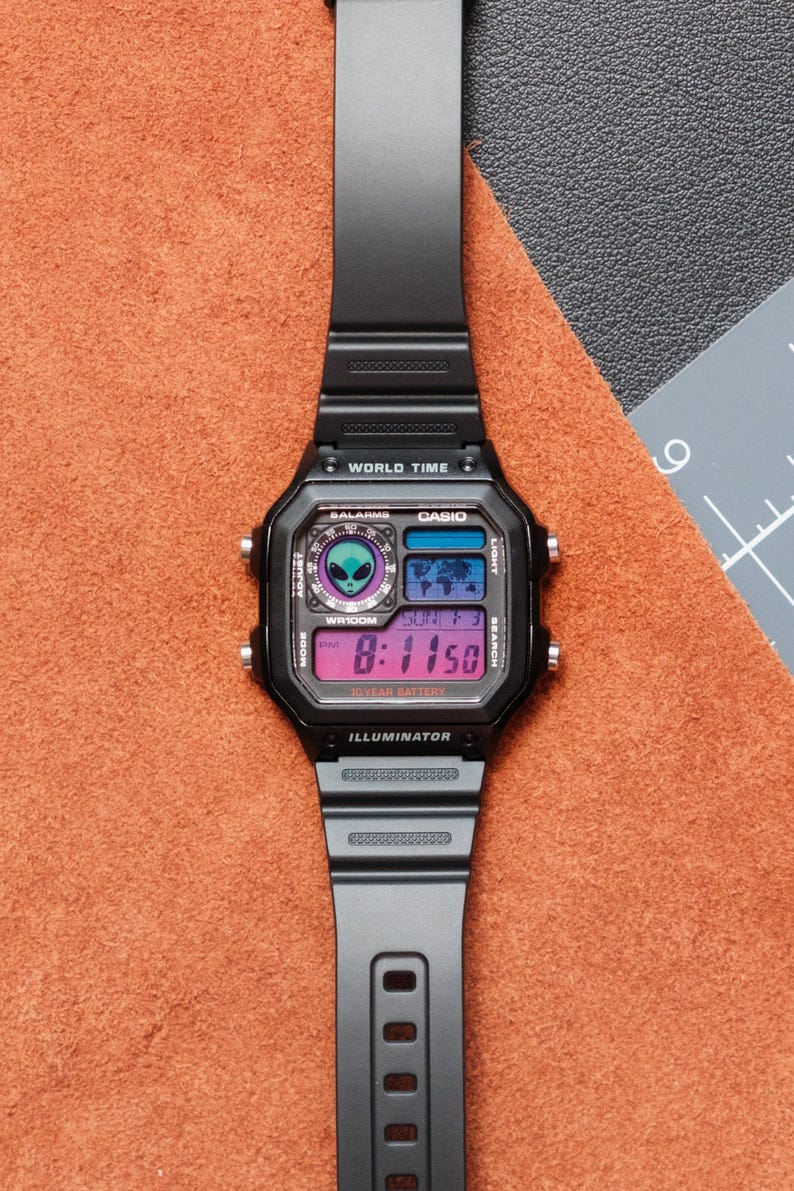 Peut inclure: Montre num&eacute;rique noire avec un cadran carr&eacute; et un bracelet noir. L'&eacute;cran affiche l'heure, la date et d'autres fonctions. La montre porte l'inscription "WORLD TIME" et "CASIO". La montre est pos&eacute;e sur une surface orange.