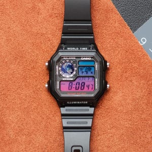 Puede incluir: Reloj digital negro con esfera rectangular y correa negra. La esfera muestra la hora, la fecha y las funciones de hora mundial. El reloj tiene el texto "CASIO" e "ILLUMINATOR" impresos en la esfera.