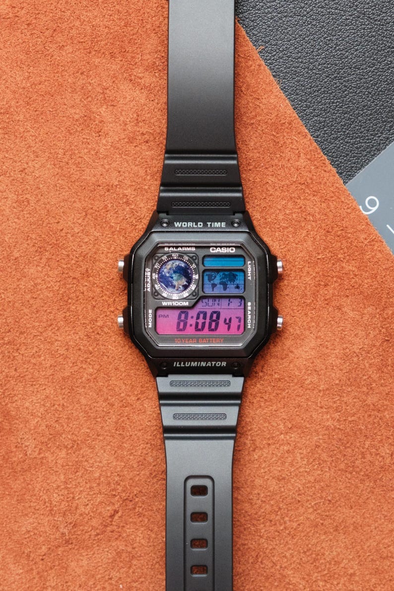 Puede incluir: Reloj digital negro con esfera rectangular y correa negra. La esfera muestra la hora, la fecha y las funciones de hora mundial. El reloj tiene el texto "CASIO" e "ILLUMINATOR" impresos en la esfera.