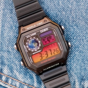 May include: Black Casio digital watch with a rectangular face displaying world time and the time 3:37:26. The watch has a black band and is set against a denim background. Features include alarms, light, and a 10-year battery.