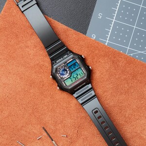 Puede incluir: Reloj digital negro con una esfera rectangular, que muestra la hora y las funciones de la hora mundial. El reloj tiene una correa negra y est&aacute; sobre una superficie marr&oacute;n. Tambi&eacute;n hay un peque&ntilde;o destornillador y tornillos.
