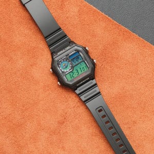 Op de afbeelding: Zwarte Casio digitale horloge met een rechthoekige wijzerplaat en een zwarte band. Het horloge geeft de tijd weer in groene cijfers en heeft het woord "Casio" op de wijzerplaat. Het horloge ligt op een oranje oppervlak.