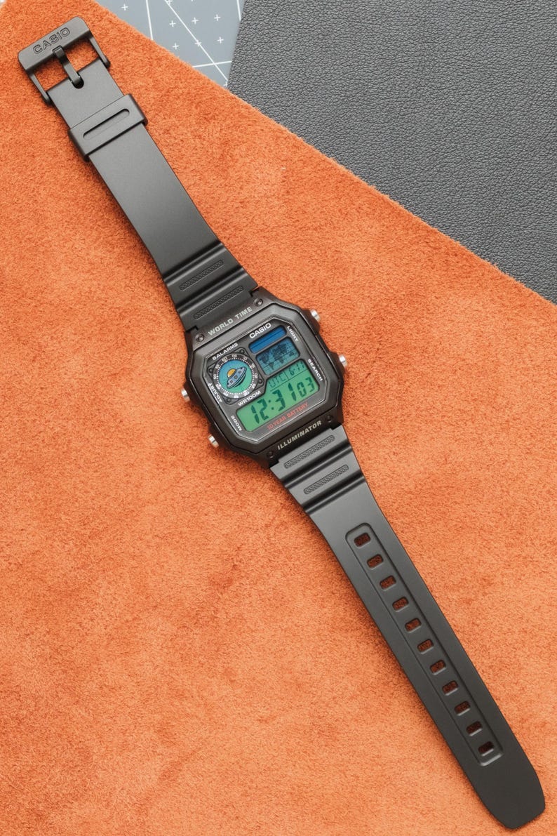 Op de afbeelding: Zwarte Casio digitale horloge met een rechthoekige wijzerplaat en een zwarte band. Het horloge geeft de tijd weer in groene cijfers en heeft het woord "Casio" op de wijzerplaat. Het horloge ligt op een oranje oppervlak.