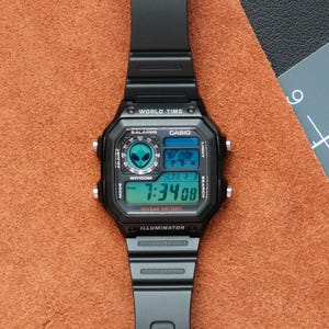 Peut inclure: Montre num&eacute;rique Casio noire avec un cadran carr&eacute; et un bracelet noir. L'&eacute;cran affiche l'heure, la date et les fonctions d'heure mondiale. La montre a un motif d'extraterrestre et le texte "WR100M" et "10 YEAR BATTERY".
