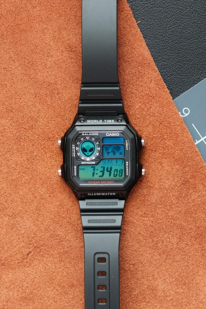 Peut inclure: Montre num&eacute;rique Casio noire avec un cadran carr&eacute; et un bracelet noir. L'&eacute;cran affiche l'heure, la date et les fonctions d'heure mondiale. La montre a un motif d'extraterrestre et le texte "WR100M" et "10 YEAR BATTERY".
