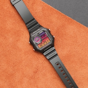 Op de afbeelding: Zwart digitaal horloge met een rechthoekige wijzerplaat en zwarte band. De wijzerplaat geeft de tijd en andere functies weer. Het horloge ligt op een oranje achtergrond.