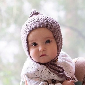 May include: A baby wearing a knitted purple bonnet with a long tassel. The bonnet has a textured pattern and covers the baby's ears.