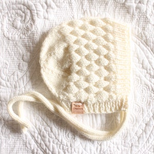May include: A white knitted baby bonnet with a textured pattern and a small leather tag that says "The Tiny Collection".