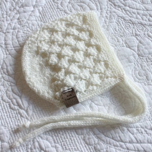 Pode incluir: Boné de bebé de malha branca com um padrão texturizado e uma etiqueta de couro marrom que diz "Kids Creations".