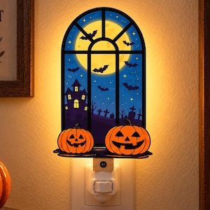May include: An arched wall plug-in night light featuring a Halloween scene with a full moon, a dark silhouette of a haunted house, a graveyard, and flying bats. Two orange jack-o'-lanterns sit at the base of the window frame, emitting a warm glow.