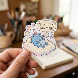 May include: A sticker featuring a blue frog wearing a party hat, lounging on a cloud, and blowing bubbles. The speech bubble says, "I require whimsy." The sticker has a pastel color scheme with a whimsical design.
