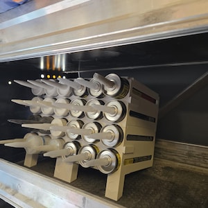 May include: A rack of caulk tubes with white nozzles. The tubes are silver and yellow, and are stored in a white plastic rack. The rack is inside a metal cabinet.