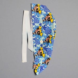 May include: A light blue scrub cap featuring a repeating pattern of blonde cartoon characters with large eyes, blue flowers, and blue hearts. The cap includes a white button on the side and a long white fabric tie for securing the head covering in place.