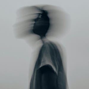 May include: A black and white photograph featuring a blurred silhouette of a figure wearing a short-sleeved shirt. The motion blur creates multiple overlapping outlines of the head and shoulders against a plain, light grey background.