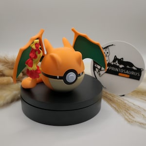 Könnte beinhalten: Eine orangefarbene und hellbraune Charizard-Figur mit grünen und gelben Flügeln, die auf einem schwarzen, kreisförmigen Ständer steht. Die Figur hat einen weißen und grauen Knopf auf dem Bauch. Ein weißer, kreisförmiger Untersetzer mit einer schwarzen Katzenillustration und dem Text "PRINTOSAURUS" befindet sich im Hintergrund.