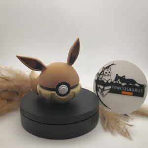 Könnte beinhalten: Eine braune und beige Eevee-Figur mit großen Ohren, einem flauschigen Schwanz und einem schwarzen Sockel. Ein weißes kreisförmiges Objekt mit einer Katzenillustration und dem Text "PRINTOSAURUS" ist ebenfalls vorhanden.