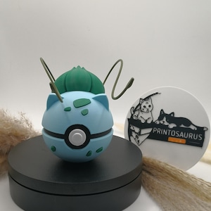 Könnte beinhalten: Eine hellblaue und grüne Bulbasaur-Pokeball-Figur mit schwarzem Sockel. Die Figur hat eine grüne Knolle oben und zwei grüne Antennen. Ein weißes und schwarzes Printosaurus-Logo ist im Hintergrund zu sehen.