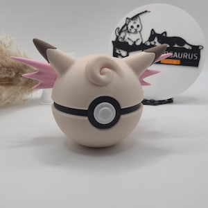 Könnte beinhalten: Eine hellbeige, runde, cartoonartige Figur mit braunen Ohren und rosa flügelartigen Akzenten. Die Figur hat ein schwarzes Band in der Mitte und einen weißen und schwarzen Kreis in der Mitte. Ein weißes kreisförmiges Objekt mit einer schwarz-weißen Katzenillustration und dem Wort "SAURUS" befindet sich im Hintergrund.