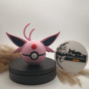 Op de afbeelding: Een roze en zwart figuur met grote oren en een rode stip op het gezicht, dat lijkt op een stripfiguur. Het figuur staat op een zwarte cirkelvormige basis. Een wit cirkelvormig bord met een kattenillustratie en de tekst "PRINTOSAURUS" is ook zichtbaar.