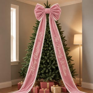 Op de afbeelding: Een grote roze fluwelen strik als piek voor in de kerstboom met twee lange linten afgewerkt met wit kant. Het linkerlint is geborduurd met Merry Christmas 2026 en het rechterlint met The Family William in wit cursief schrift.