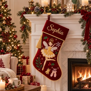 May include: A deep red velvet Christmas stocking featuring the embroidered name Sophia in gold script. The stocking is decorated with a fairy in a yellow dress, a gold bell, pink ballet slippers, and green holly leaves with red berries.
