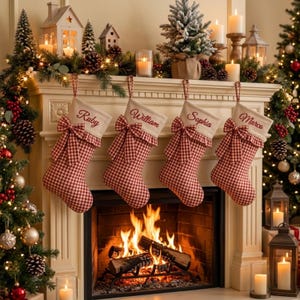 May include: Four red and white gingham Christmas stockings hanging from a fireplace mantel. Each stocking features a ruffled top with a bow and is embroidered with the names Ruby, William, Sophia, and Marco. The mantel is decorated with pine garlands, miniature houses, candles, and small trees.
