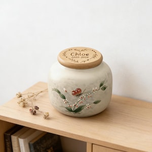 Op de afbeelding: Een keramische urn met een houten deksel. De urn is gebroken wit met een geschilderd bloemmotief en een rode vlinder. Het houten deksel is gegraveerd met de naam "Chloe" en de jaren "2020-2026".