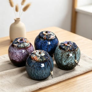 May include: Four ceramic jars with lids. The jars are in shades of blue, purple, and green with speckled patterns. Each jar has a fabric lid with a floral design and a drawstring closure. A vase with dried flowers is in the background.