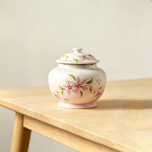 Könnte beinhalten: Ein kleiner, runder Keramiktopf mit Deckel und rosa Blumenmuster. Der Topf ist cremefarben mit rosa Blüten und grünen Blättern. Der Deckel hat ein passendes Blumenmuster. Der Topf steht auf einer hellen Holzoberfläche.