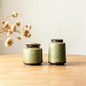 Puede incluir: Dos tarros de cerámica verde con tapas de madera sobre una superficie de madera. Los tarros tienen un esmalte moteado y una base marrón oscuro. Una tapa está ligeramente entreabierta, revelando un interior oscuro. Flores secas en el fondo.
