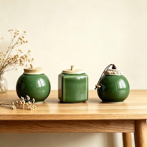 May include: Three green ceramic jars with wooden lids and a drawstring closure on a wooden table. The jars are different shapes: round, square, and spherical. The jars are a glossy green color and have a smooth finish. Decorative dried flowers are in the background.
