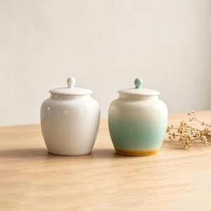 May include: Two ceramic jars with lids. One is solid white, the other has a gradient of white to light green, with a yellow base. Both jars have a small knob on top of the lid. They sit on a wooden surface.