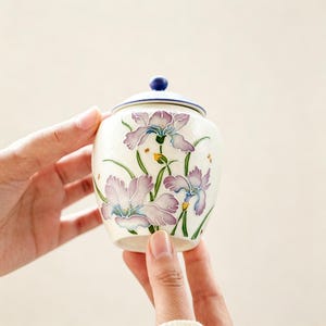 May include: A small, cream-coloured ceramic jar with a lid, decorated with purple and blue iris flowers and green leaves. The lid is blue with a small knob. The jar is held by two hands.