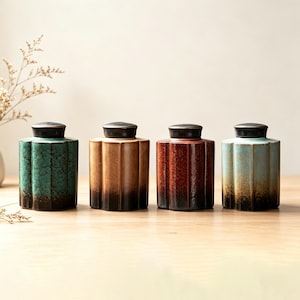 Może przedstawiać: Cztery ceramiczne słoiki z pokrywkami. Każdy słoik ma unikalny kolor: morski, brązowy, czerwony i niebieski. Słoiki mają żebrowany wzór i ciemnobrązową pokrywkę. Słoiki stoją na jasnobrązowej drewnianej powierzchni.