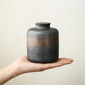 Peut inclure: Un pot en céramique fait à la main, avec une base gris foncé et une bande de glaçure couleur bronze. Le pot a un corps arrondi et un petit couvercle plat. Le pot est tenu dans la main d'une personne sur un fond neutre.