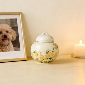 Op de afbeelding: Een crèmekleurige keramische urn met een deksel, versierd met handgeschilderde gele madeliefjes en groene bladeren. Een ingelijste foto van een hond en een brandende kaars staan op de achtergrond.