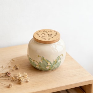 May include: A round, cream-coloured ceramic jar with a light wood lid. The lid features an engraved heart design with the text "Chloe 2020-2026". The jar is decorated with a relief pattern of green leaves and white lily of the valley flowers.