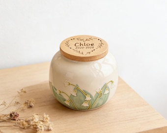 May include: A round, cream-colored ceramic jar with a light wood lid. The lid features an engraved heart design with the text "Chloe 2020-2026". The jar is decorated with a relief pattern of green leaves and white lily of the valley flowers.