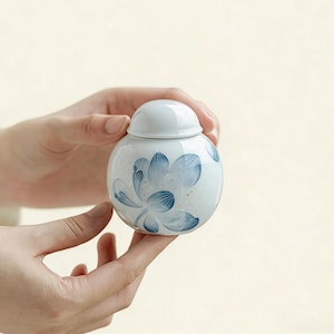 Puede incluir: Un pequeño tarro redondo de cerámica blanca con tapa, decorado con motivos florales azules. El tarro se sostiene con dos manos, mostrando su delicado diseño. El tarro mide aproximadamente 7.6 cm de diámetro, perfecto para guardar objetos pequeños.